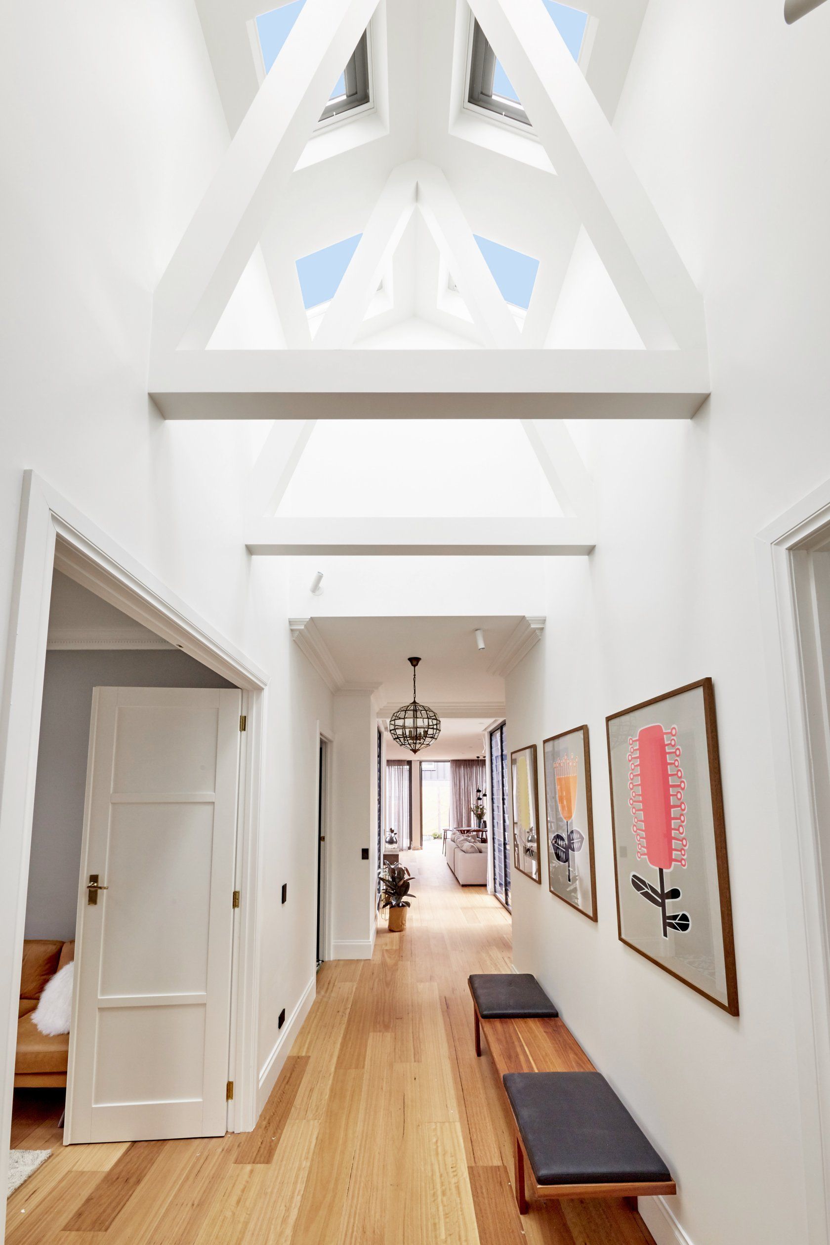 Long hallway with light wood floors and white walls, benches, artwork, and a high ceiling with skylights.
