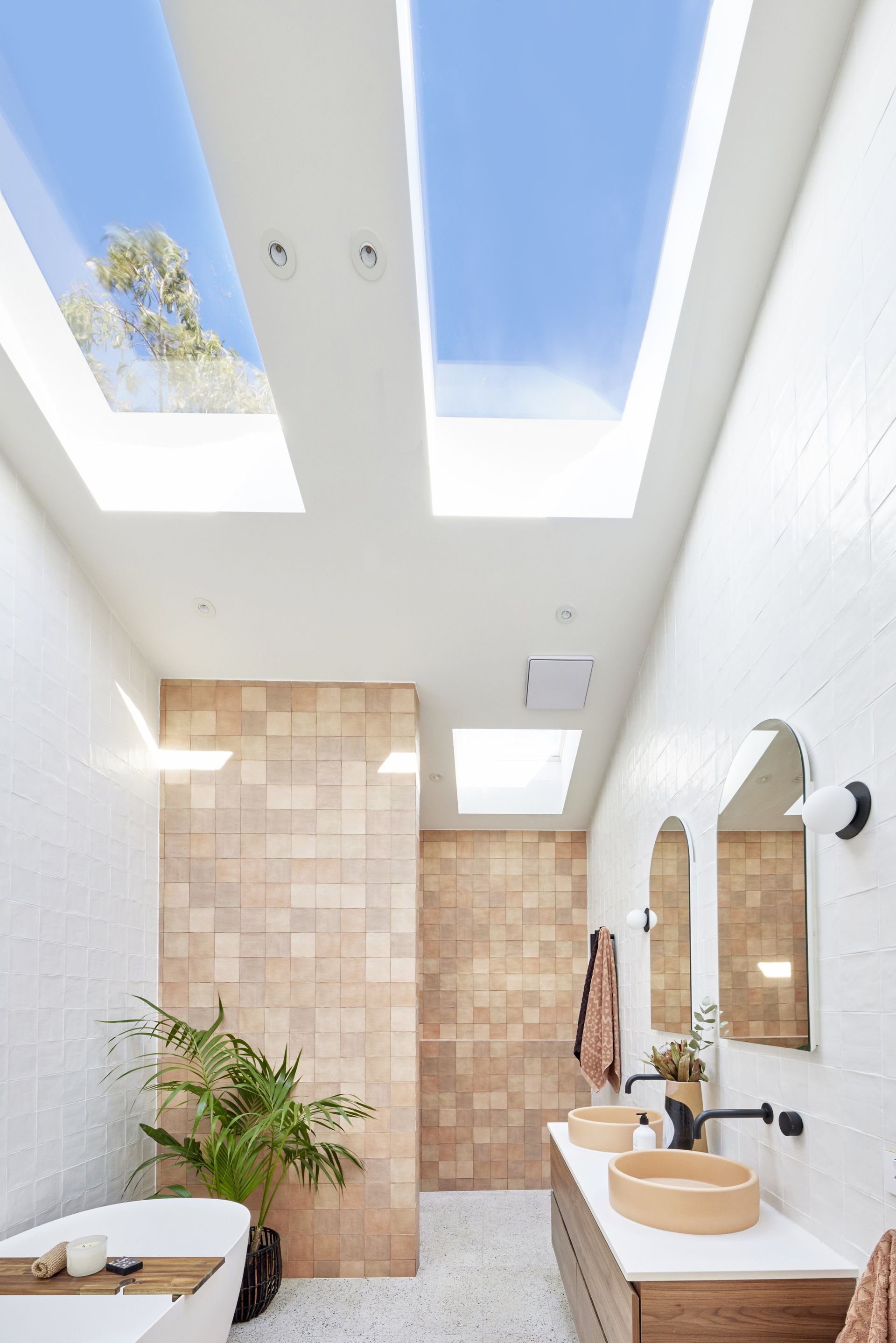 Bathroom with skylights, beige tiles, white walls, wooden vanity, and two round sinks.