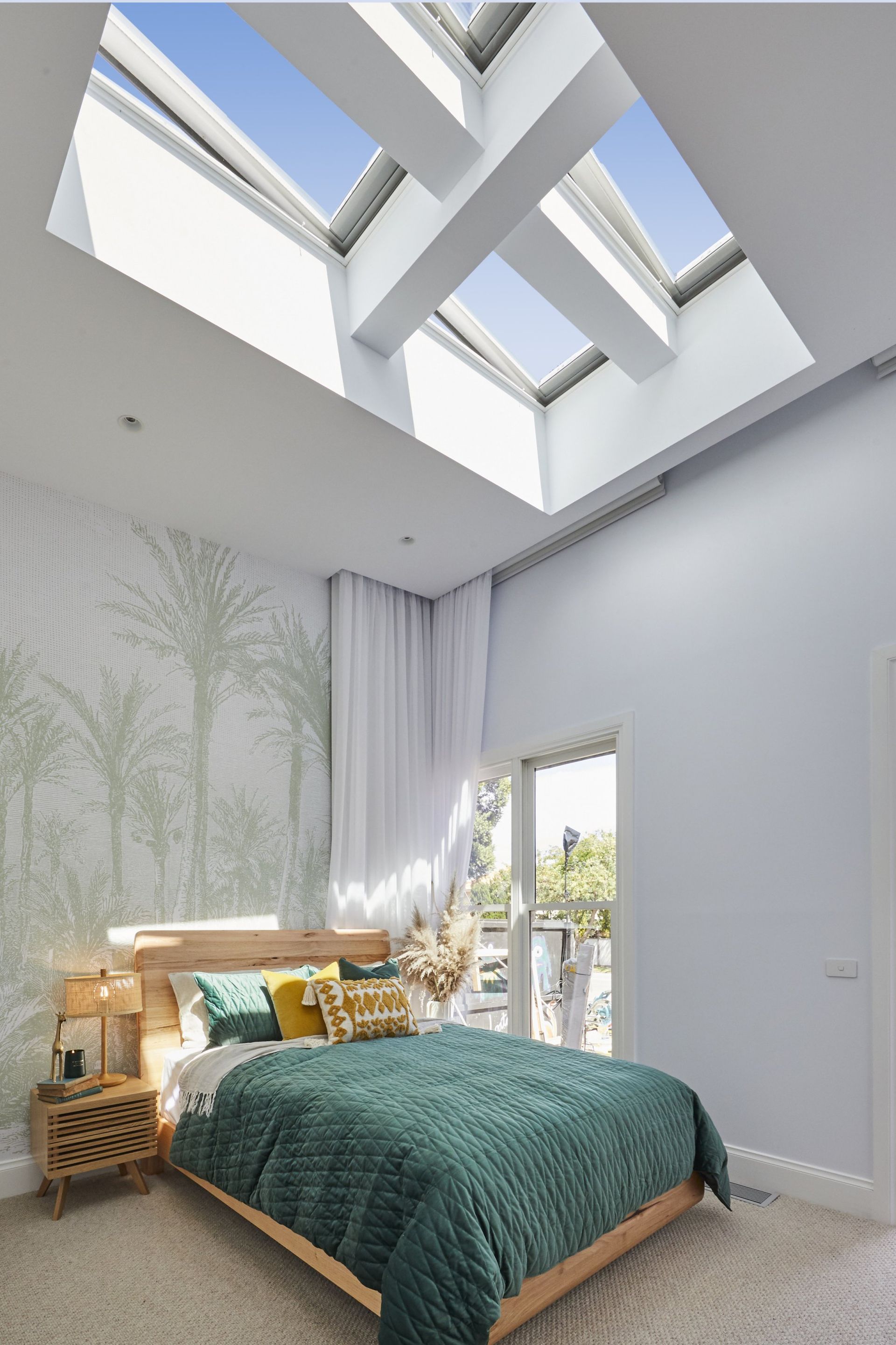 Bedroom with a skylight, bed with teal quilt, palm tree wallpaper, and a wooden bedside table.