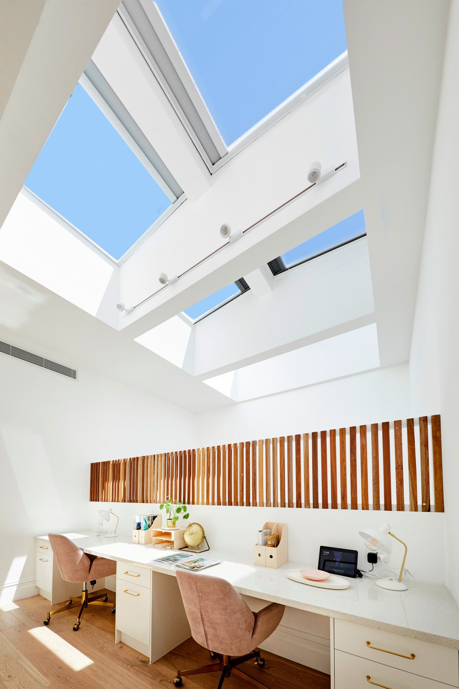 Bright white home office with desk under skylights, featuring two pink chairs, wood accent, and clear blue sky.
