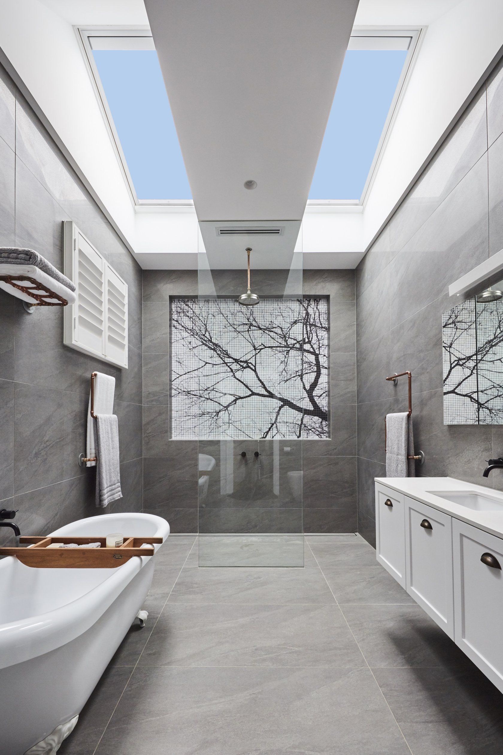 Modern bathroom with skylight, gray tile, bathtub, shower with tree art, and white vanity.
