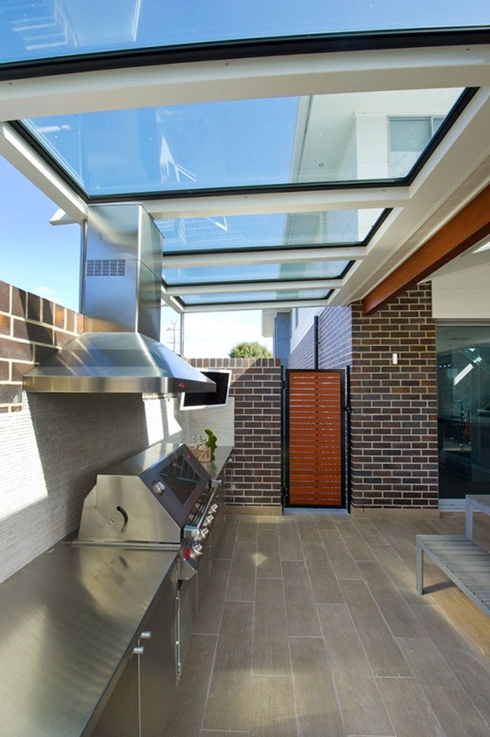 Outdoor kitchen with stainless steel appliances, brick walls, glass roof, and wooden screen.