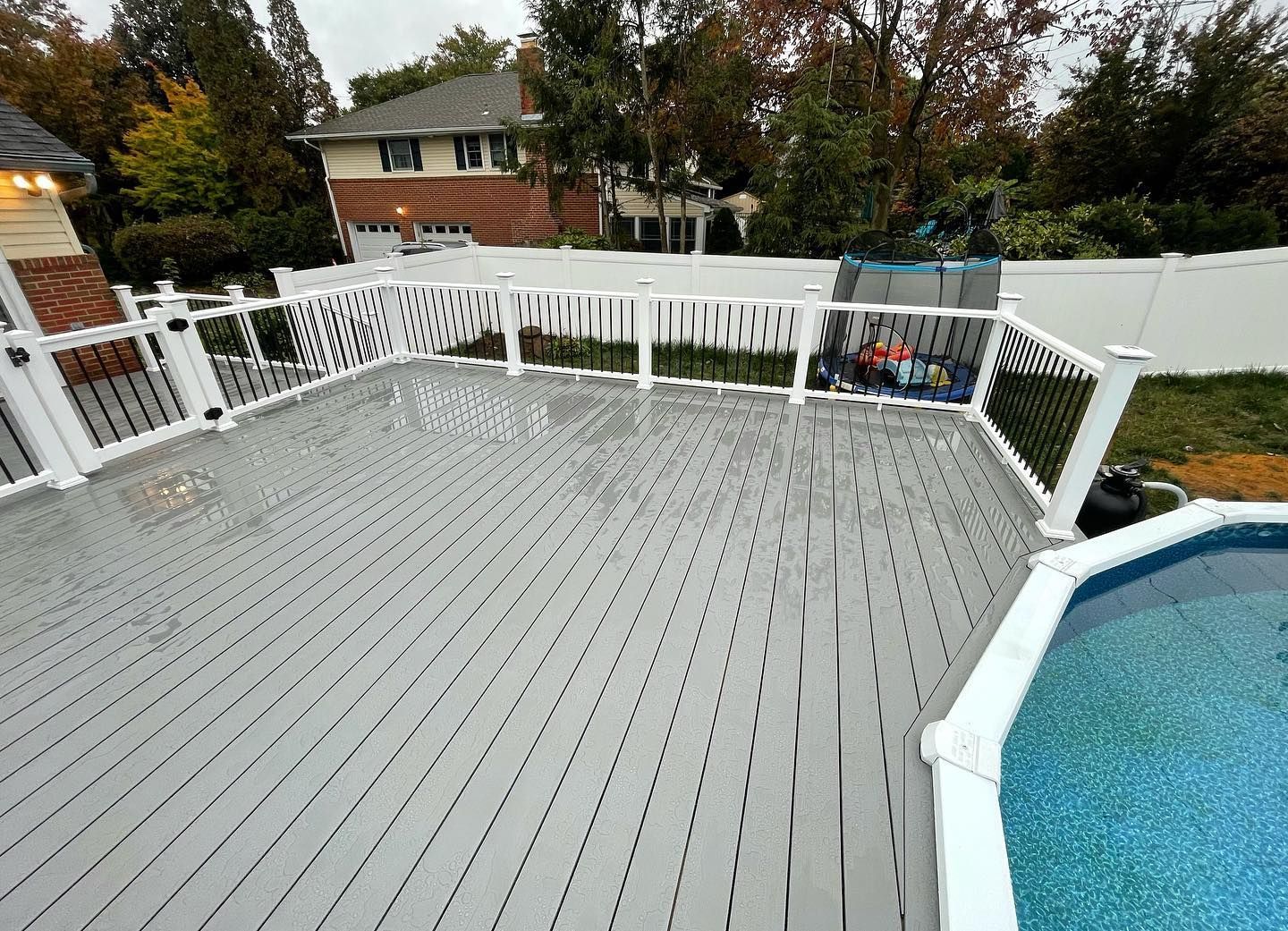 Grey wooden deck with white railing beside a pool. House in the background. Overcast day.