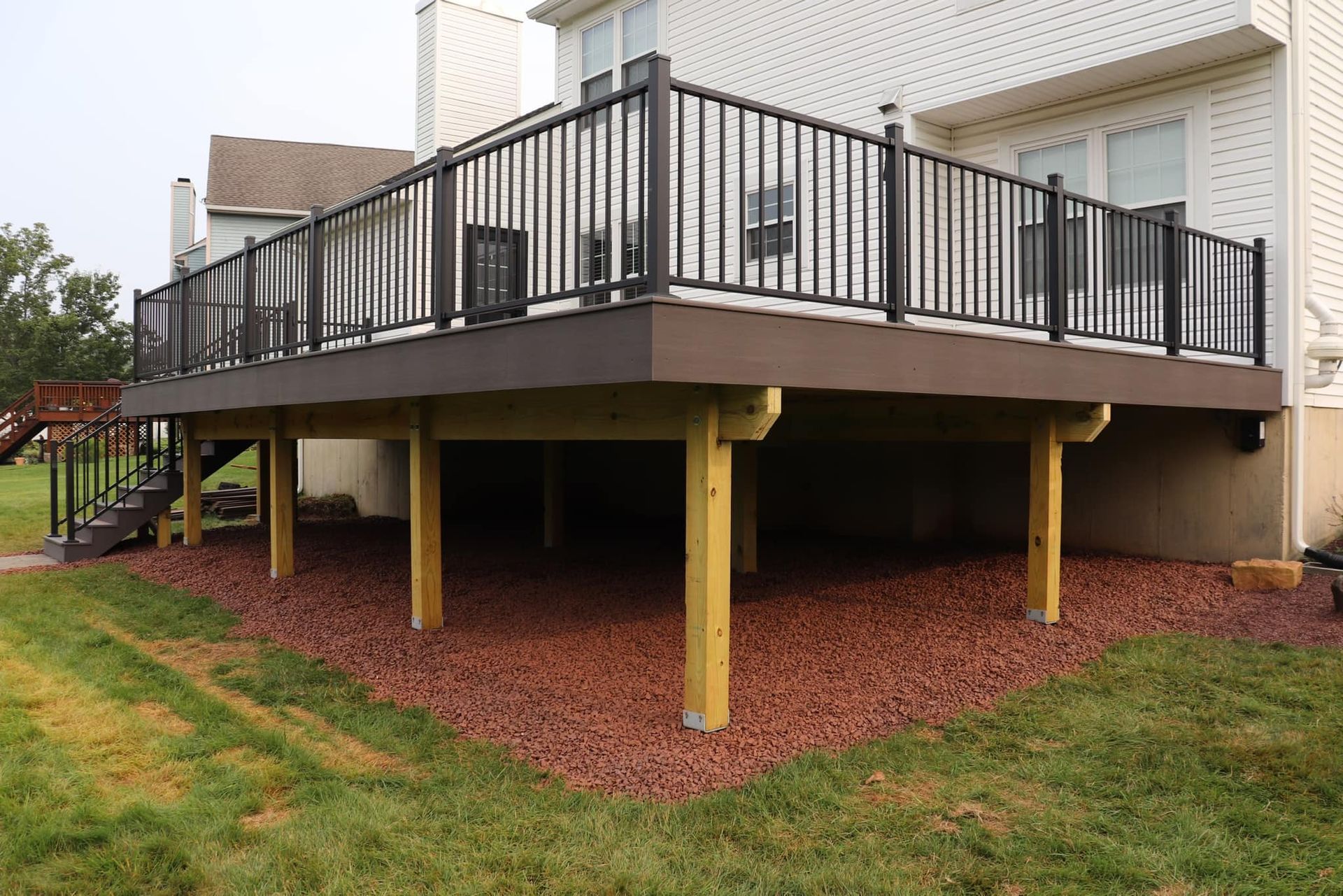 Elevated deck with black railing and brown composite decking; supported by wooden posts and surrounded by red gravel.
