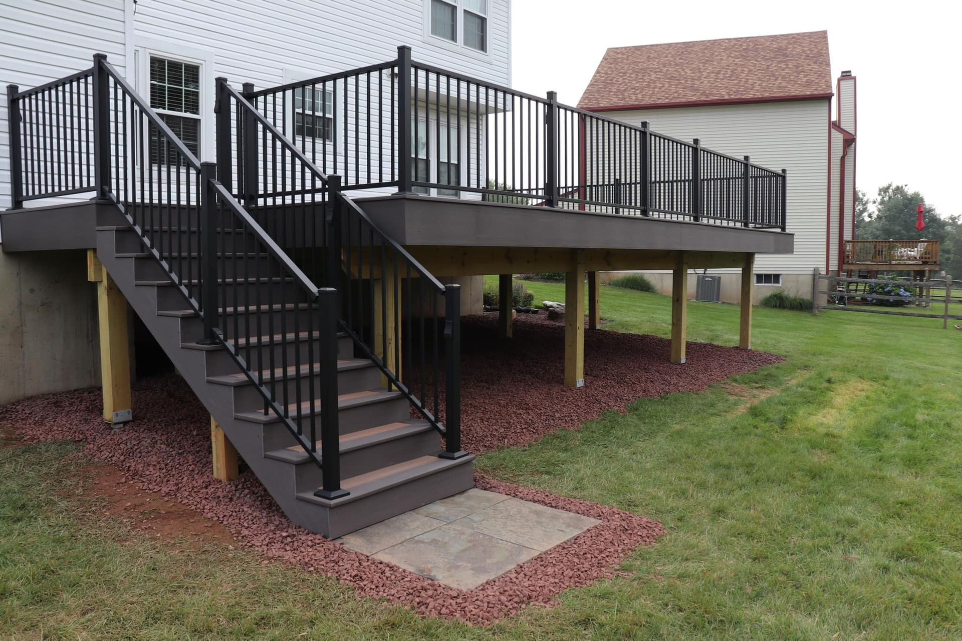Black railing surrounds a deck with stairs. Brown deck boards and tan support posts. Reddish-brown gravel around the base.