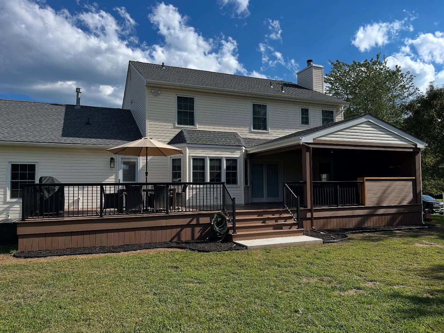 Backyard with a two-story beige house, deck, and covered patio on a sunny day. Brown deck, black railings, and a green lawn.