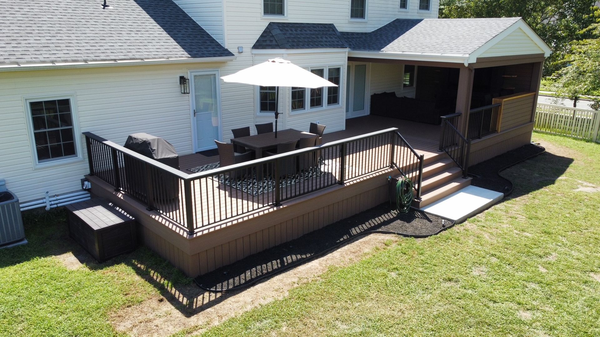 Backyard deck with furniture and umbrella attached to a house with white siding and a brown screened-in porch.