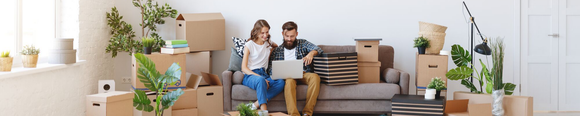 Two people sitting on a gray couch in a bright living room with moving boxes and houseplants