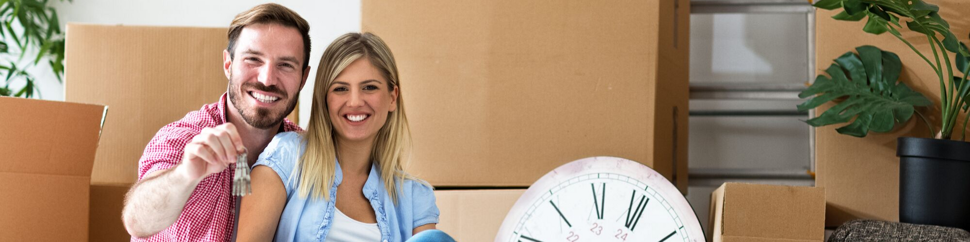 Smiling couple in a kitchen holding a pink piggy bank beside a large clock
