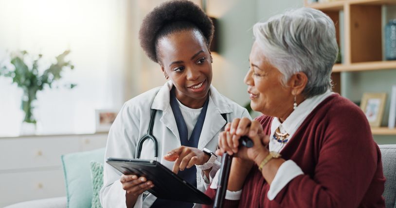 A doctor shows an elderly patient a tablet in a home setting. Both are smiling.