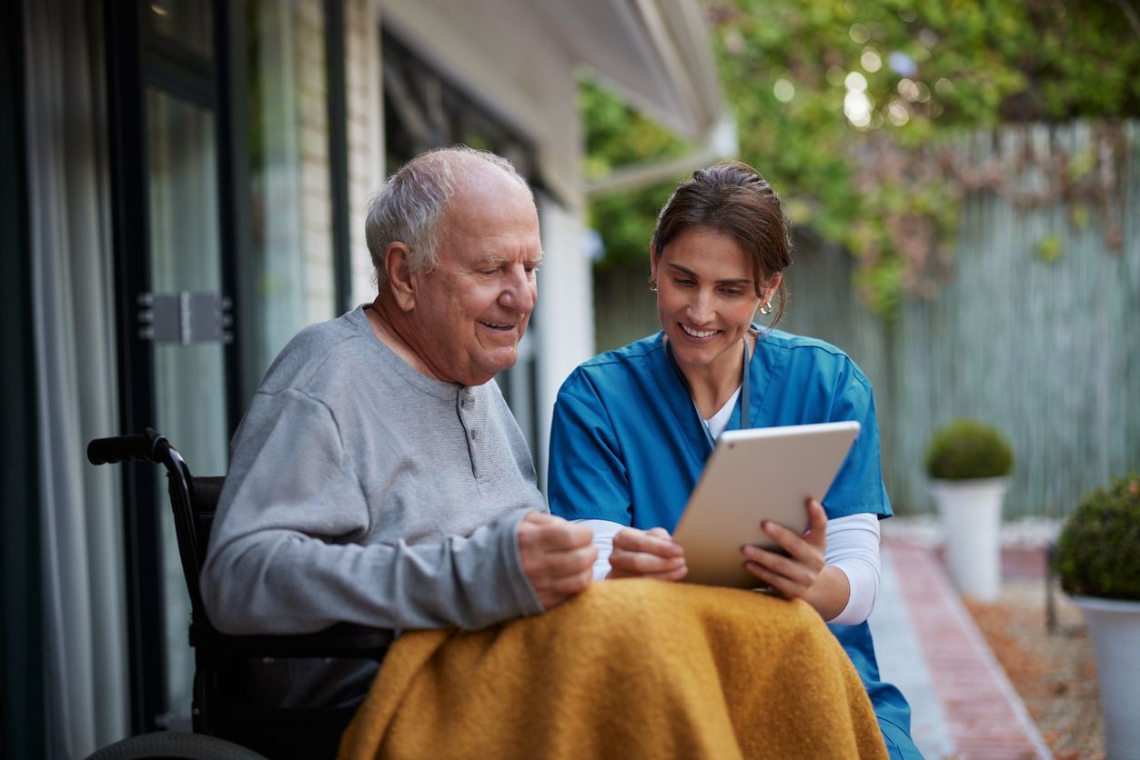 An older person in a wheelchair and a caregiver looking at a tablet together outdoors.