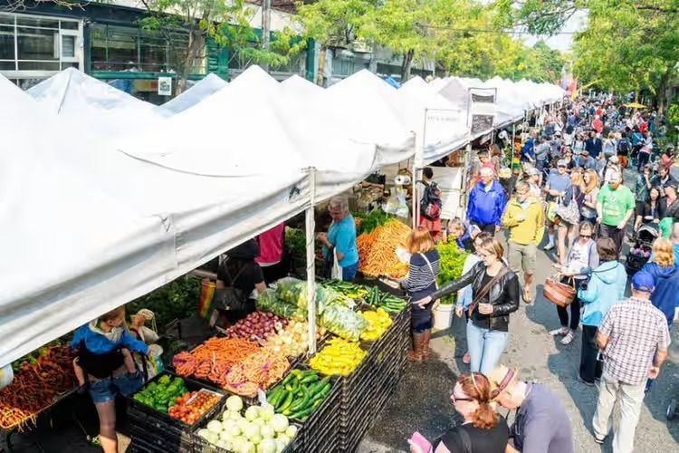 Crowded outdoor market with white tents, colorful produce stands, and shoppers walking along the aisle