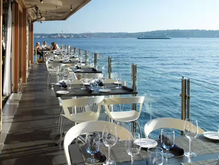 Outdoor waterfront dining deck with white chairs and tables overlooking calm blue water