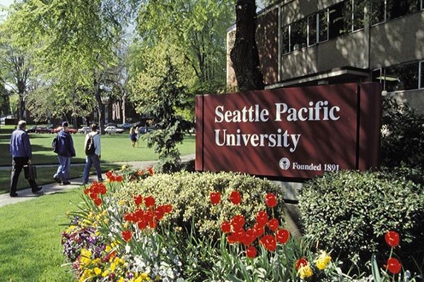 Seattle Pacific University sign by flower bed on a sunny campus lawn, with people walking nearby