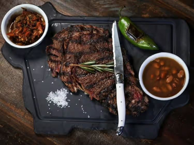 Grilled steak with rosemary, jalapeño, and two side bowls on a black tray with a knife and salt on a wooden table
