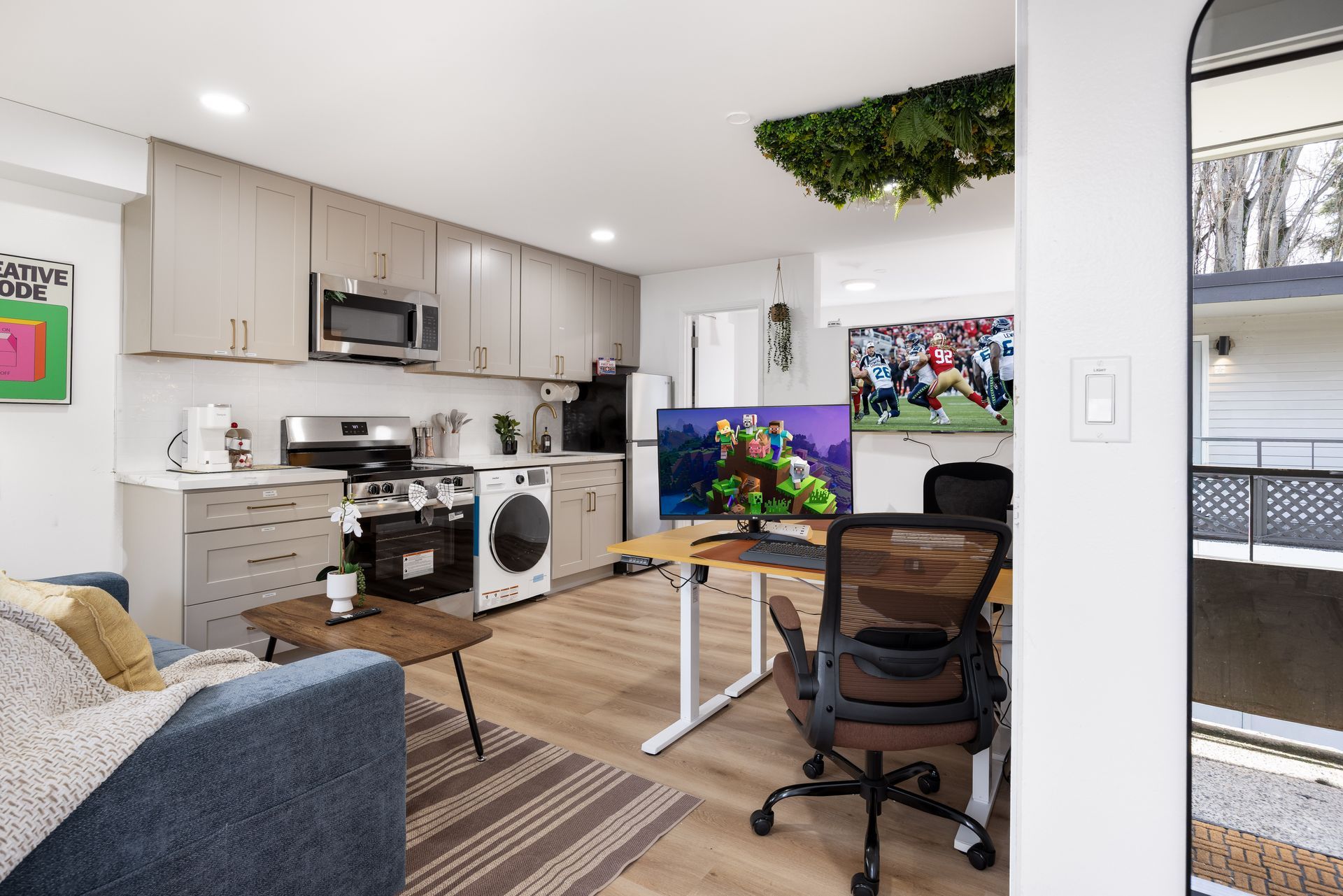 Home office and kitchen with a desk, computer monitor, sofa, and laundry area in a bright open space