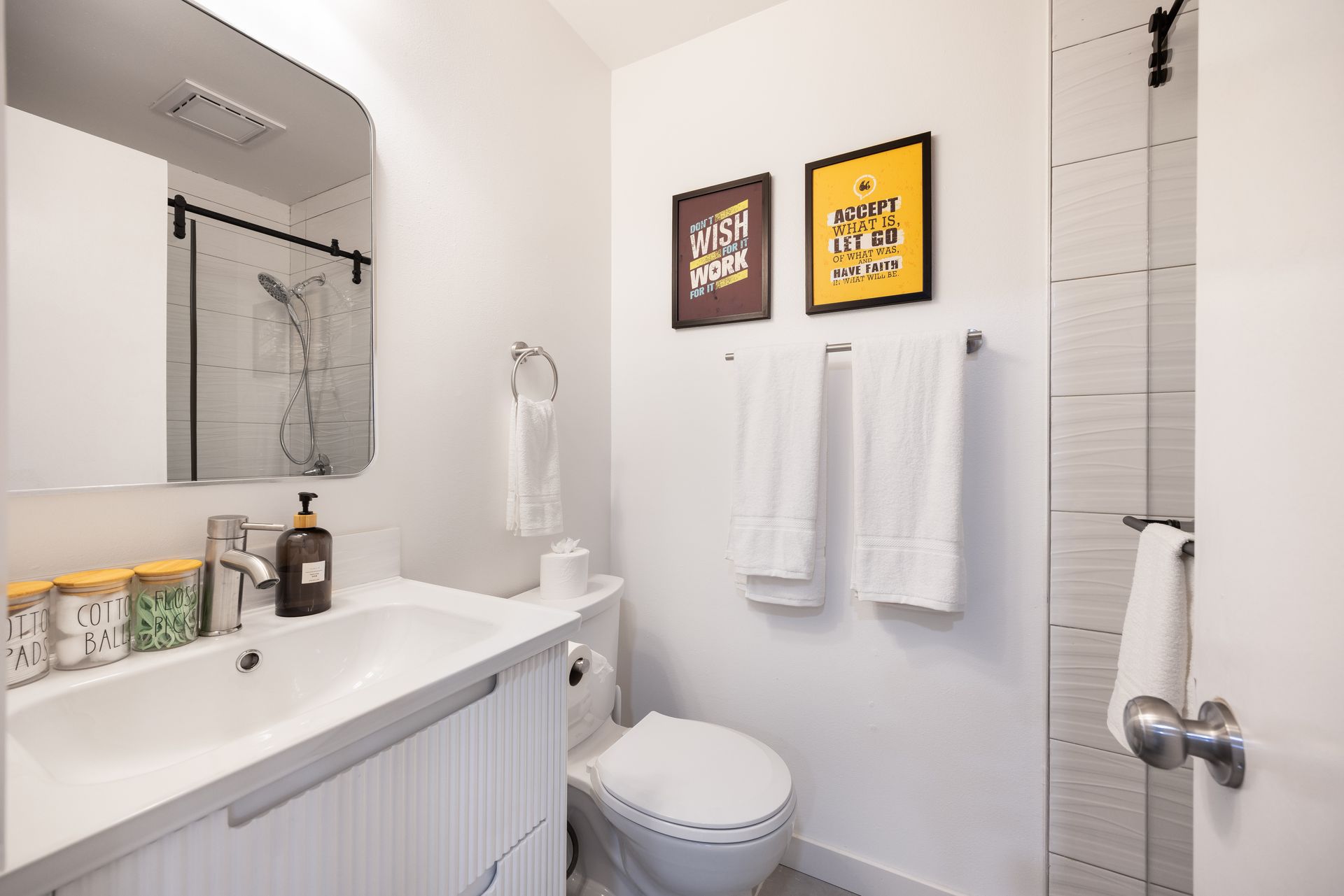 Bright bathroom with white vanity, mirror, toilet, shower, and two framed prints on the wall.