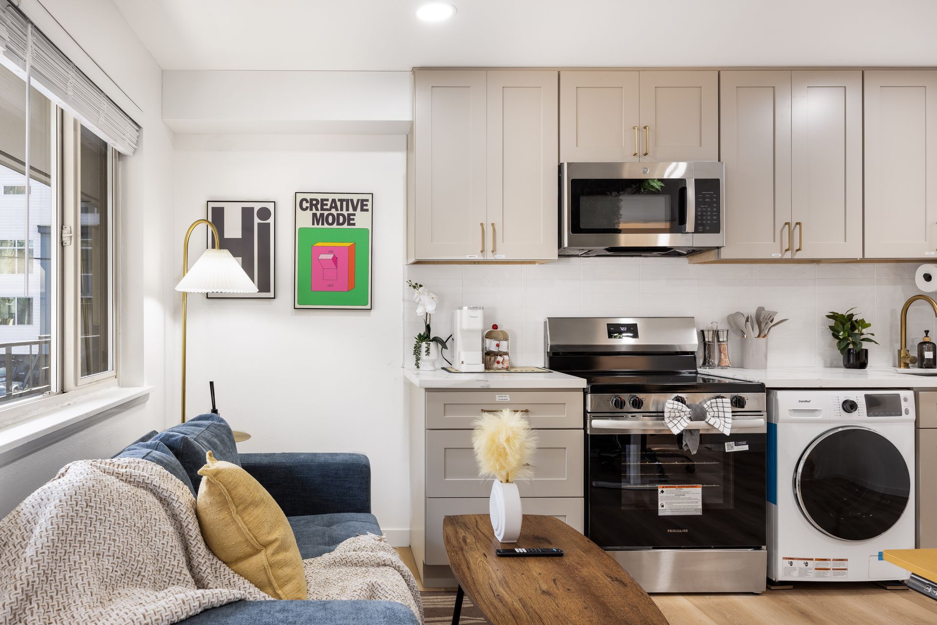 Bright modern kitchen and living room with white cabinets, stainless appliances, and a sofa by the window.