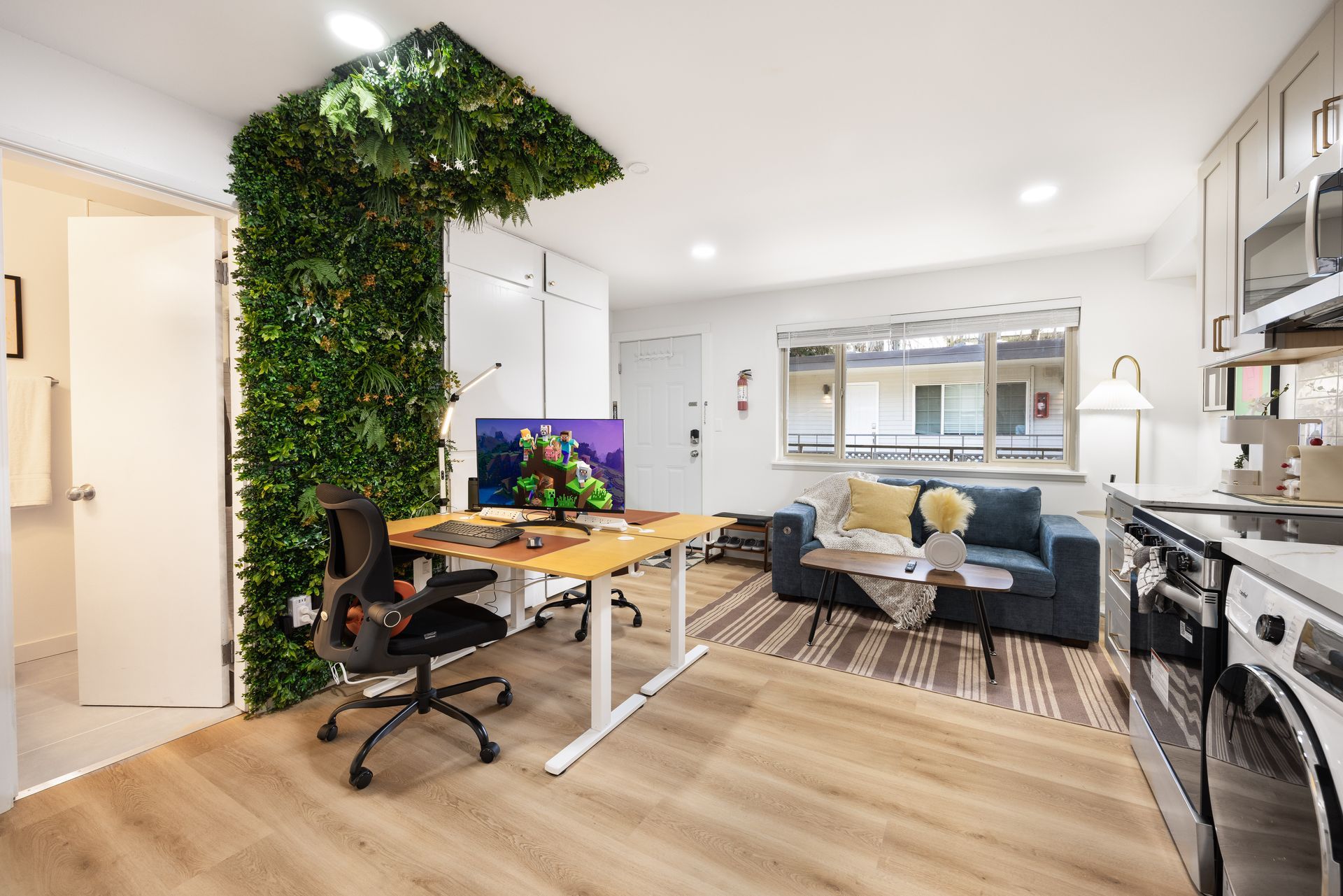 Modern open-plan home office and living room with green plant wall, desk, sofa, and kitchen in the background