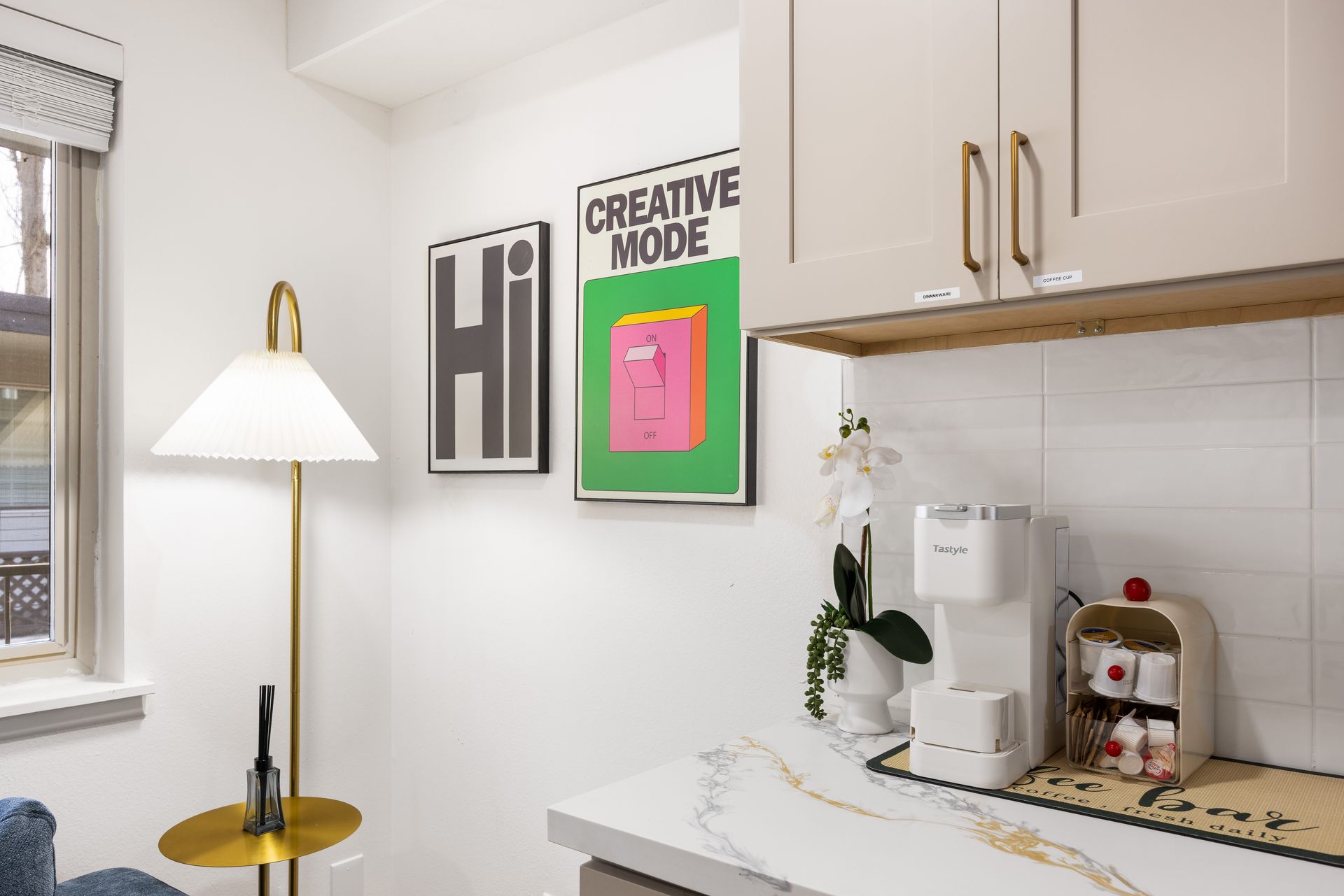 Bright kitchen corner with framed art, white cabinets, coffee machine, and a small lamp by the window.