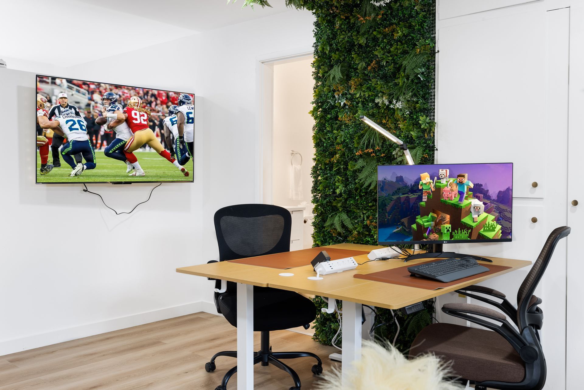 Modern office with wall-mounted TV, desk, dual monitors, and black chairs beside a green plant wall
