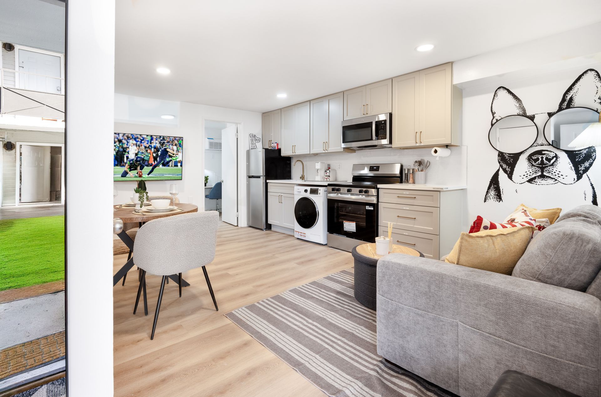 Bright modern living room and kitchen with gray sofa, dining table, white cabinets, and striped rug