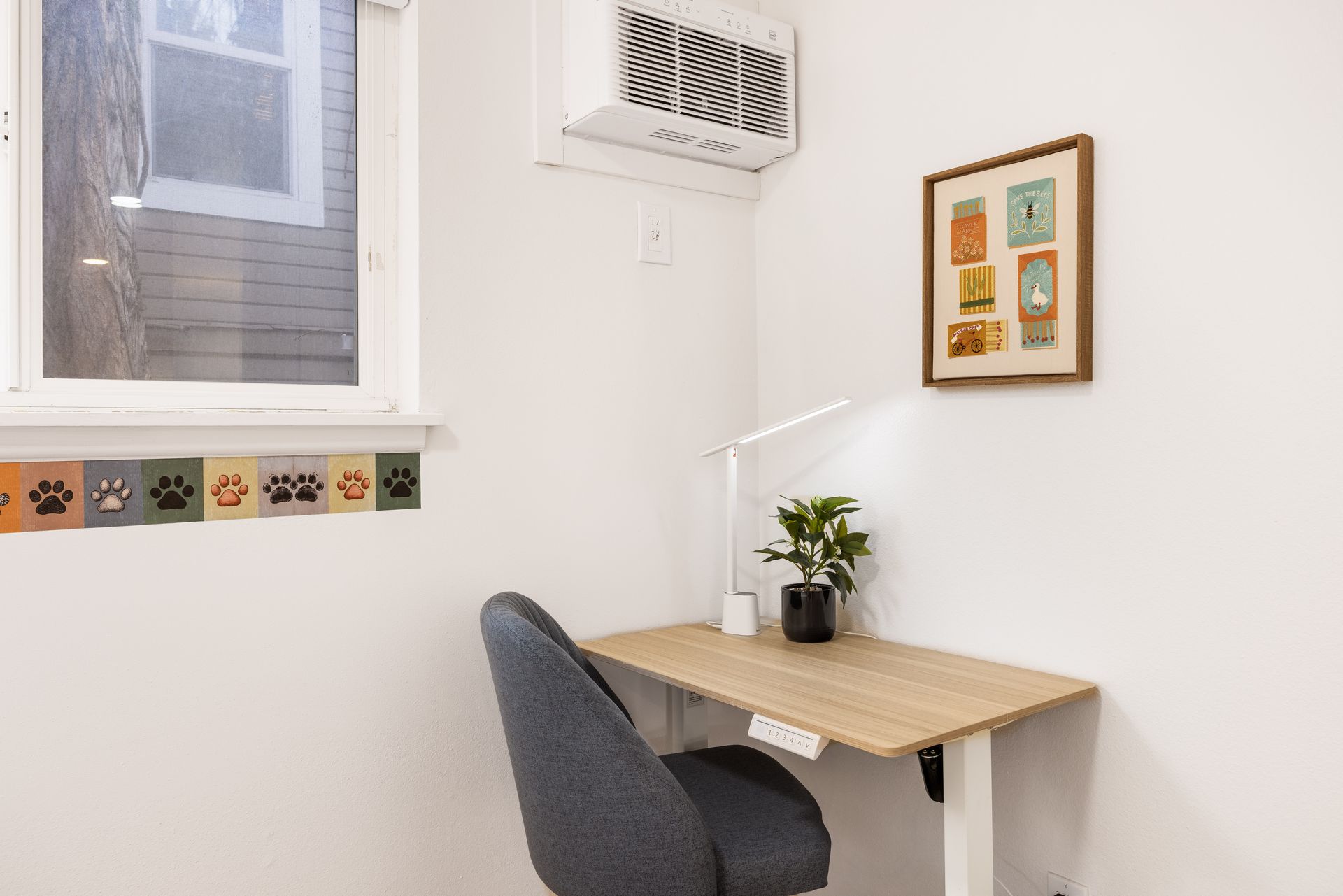 Small white office nook with desk, gray chair, plant, lamp, wall art, and a window