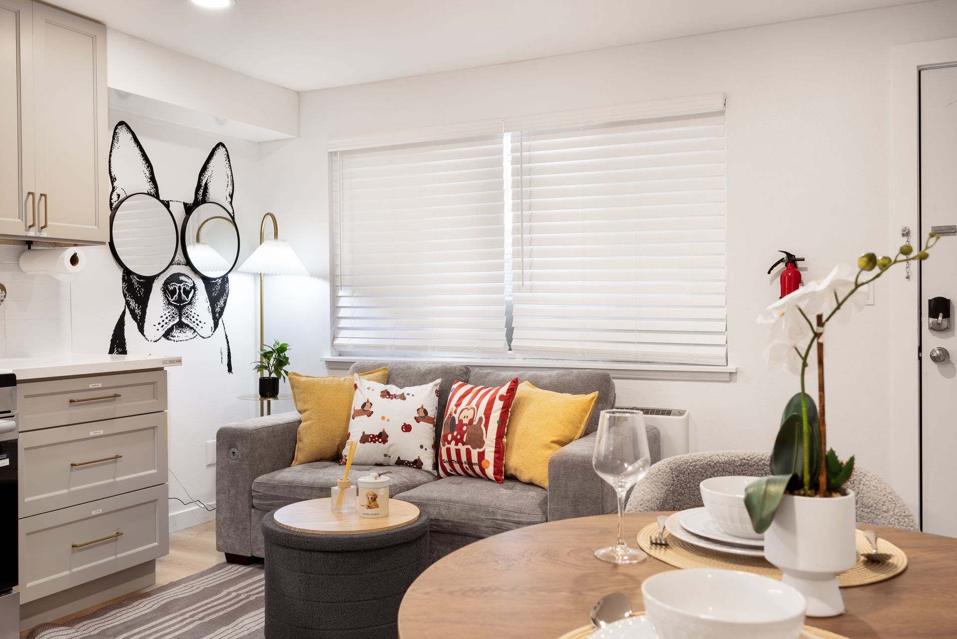 Bright modern living room with gray sectional sofa, patterned pillows, white blinds, and decorative wall mirror