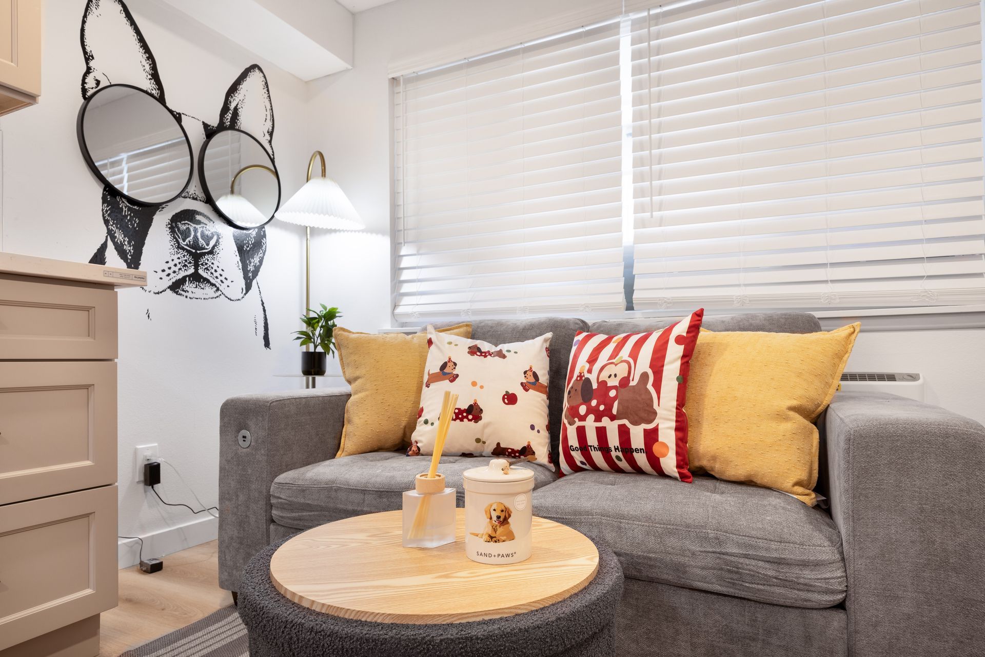 Cozy living room with gray sofa, yellow and patterned pillows, round coffee table, and wall mirrors near blinds
