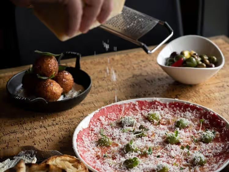 Hand grating cheese over a pink pasta dish with bowls of fried balls and olives on a wooden table
