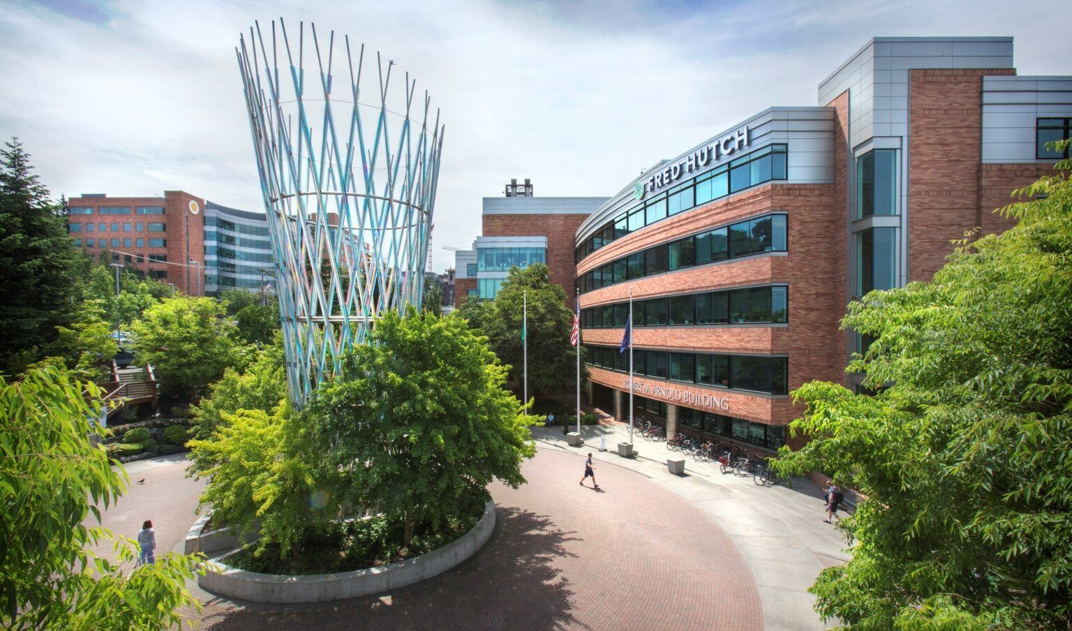 Modern campus courtyard with brick buildings, trees, a curved path, and a glass tower sculpture