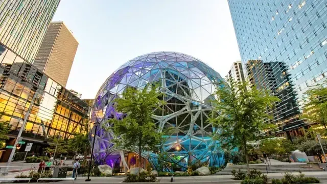 Glassy geodesic dome in a modern plaza, framed by tall buildings and trees, with colorful reflections.