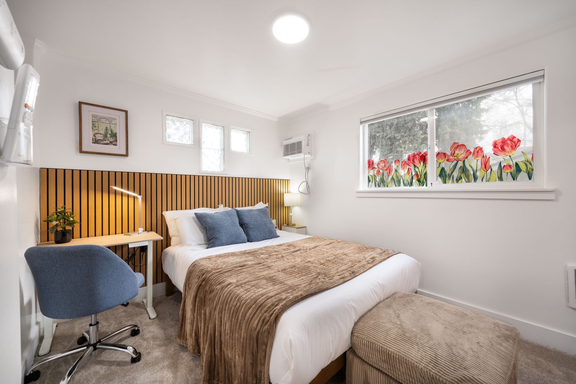 Bright bedroom with a bed, desk, blue chair, striped headboard, and floral window view