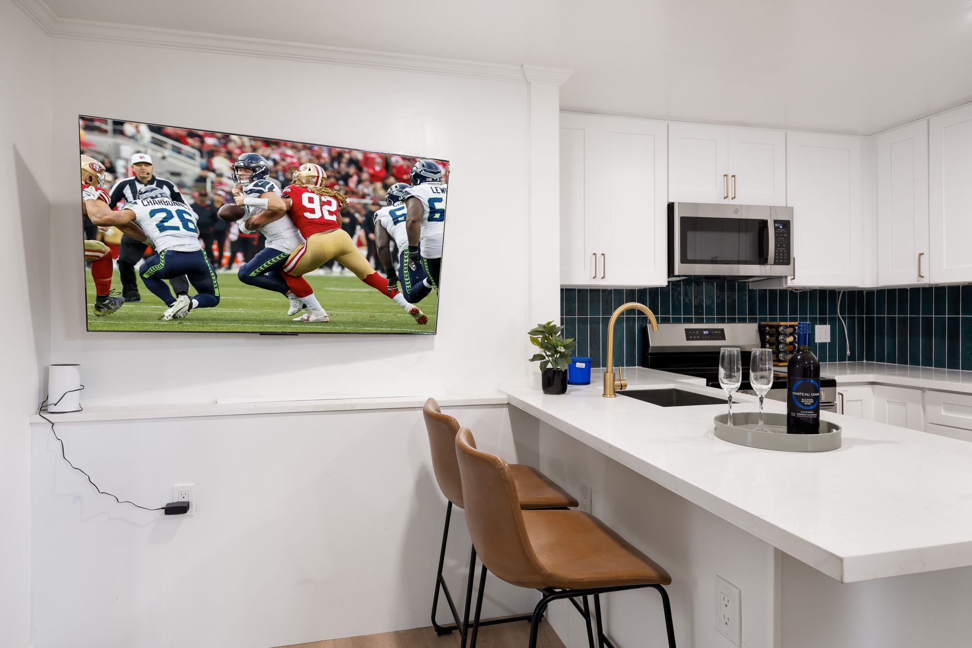 Modern white kitchen with island, brown chairs, wall-mounted TV showing a football game