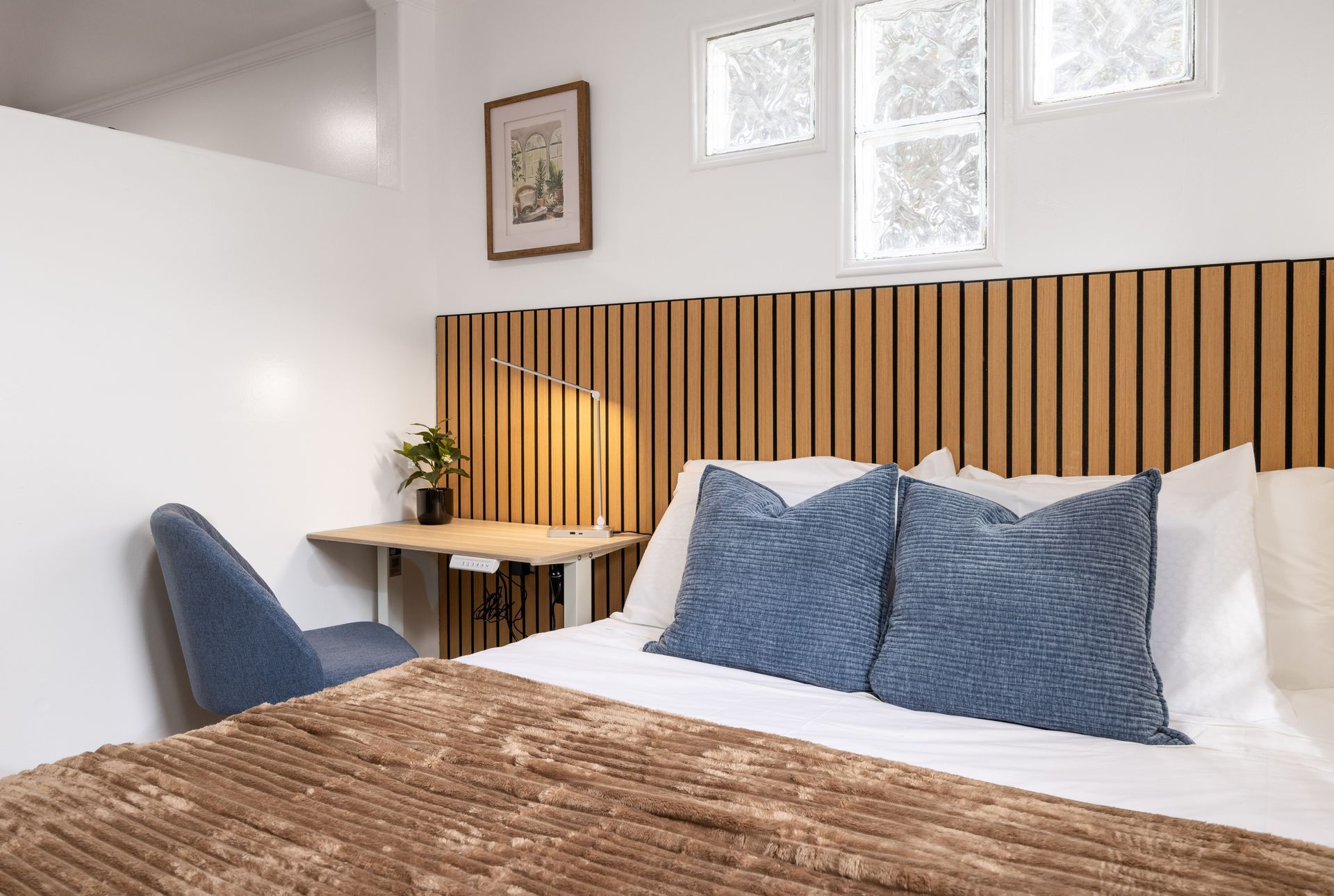 Modern bedroom with blue pillows, wooden slat headboard, desk, chair, and bright windows