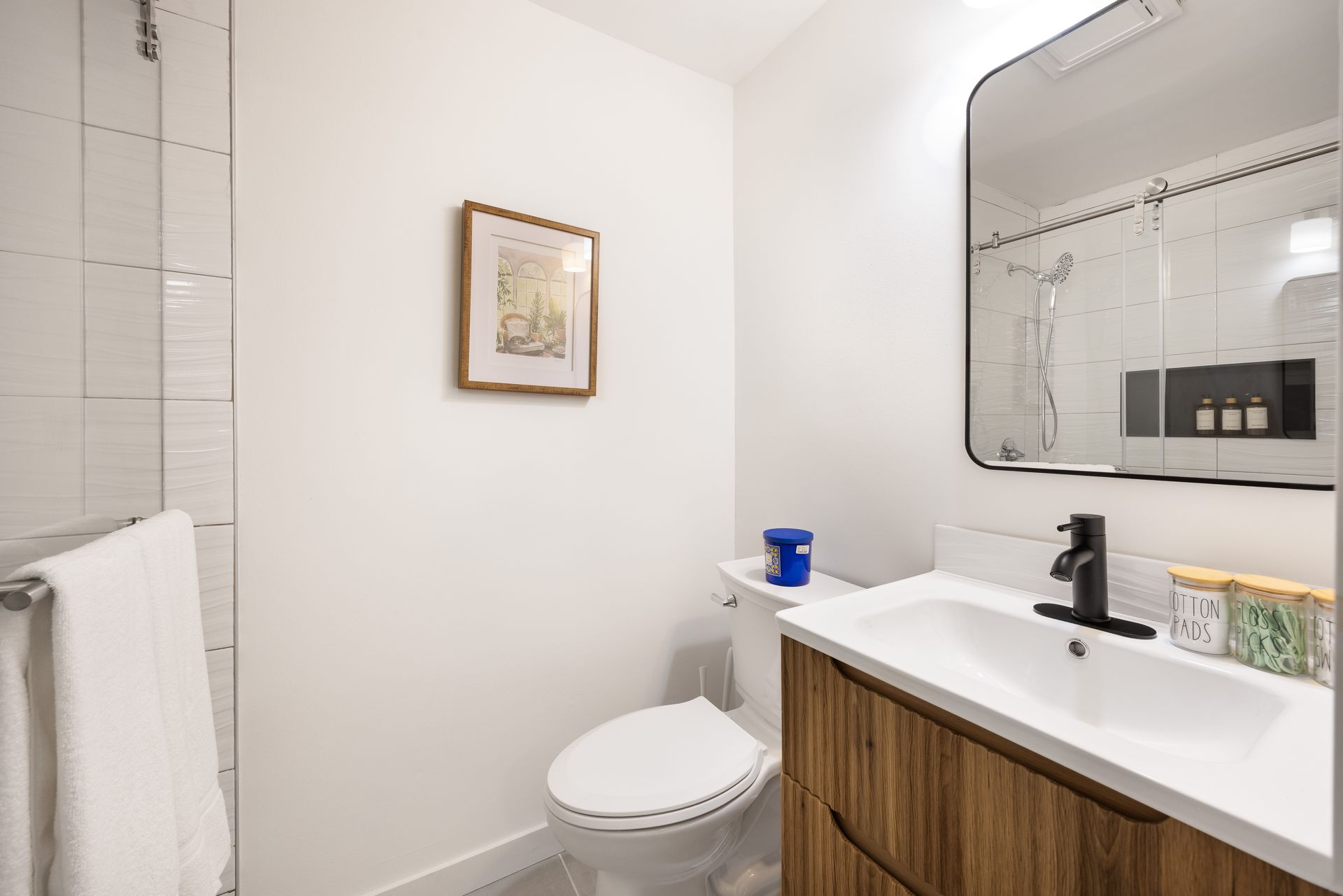 Compact white bathroom with toilet, wood vanity, black faucet, and framed wall art