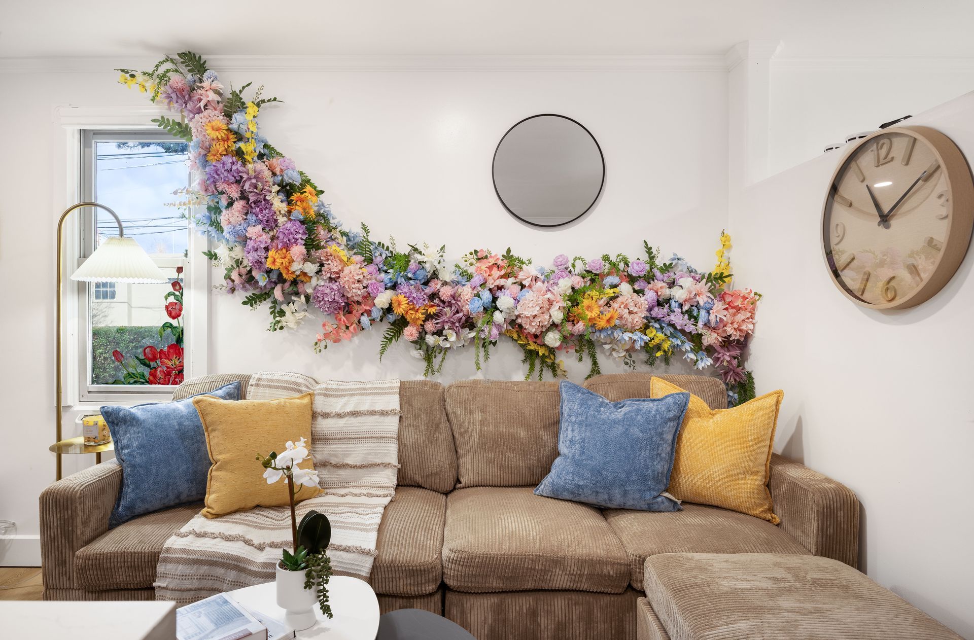 Cozy living room with tan sofa, blue and yellow pillows, and colorful floral garland on the wall