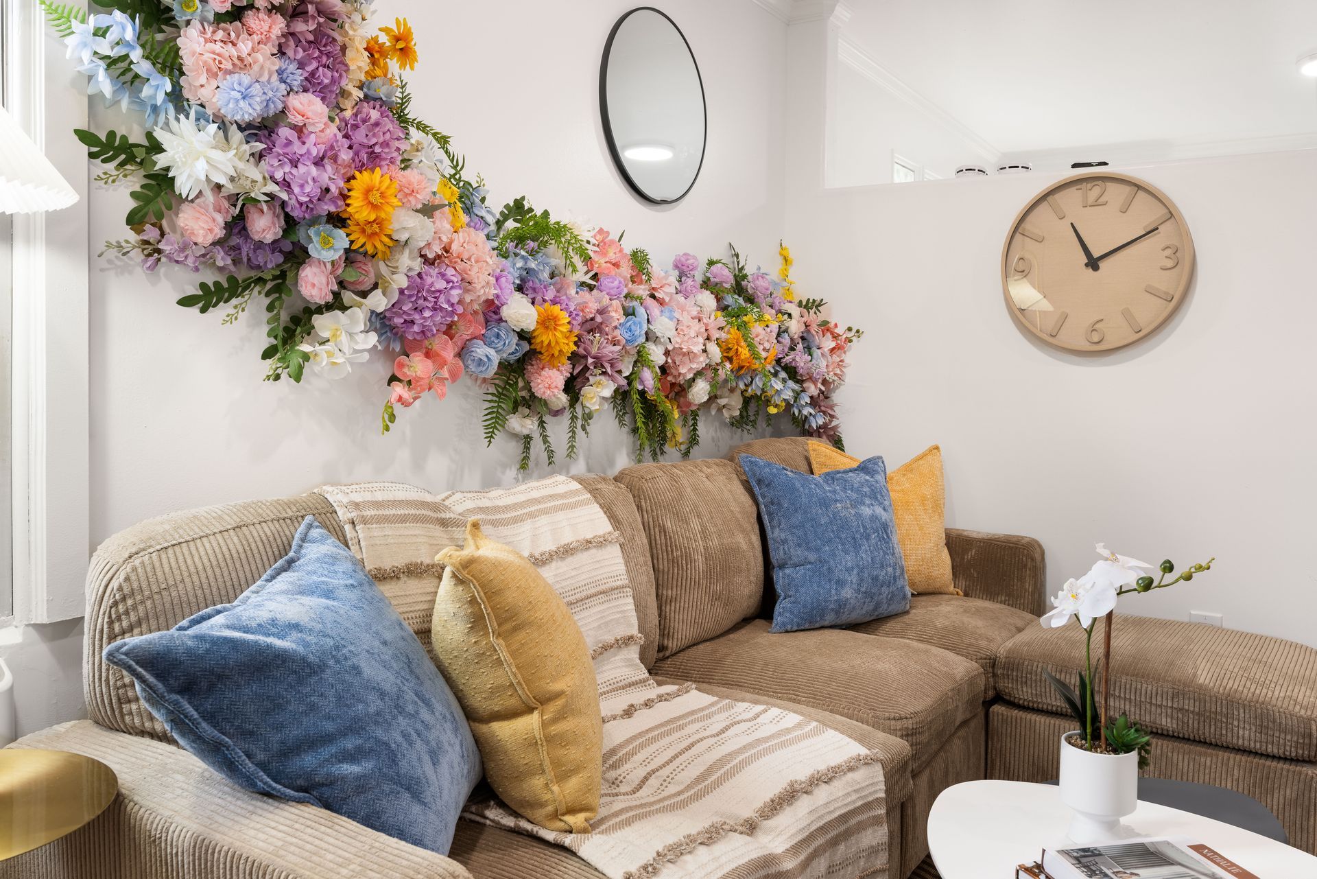 Cozy living room with beige sofa, blue and gold pillows, floral wall arrangement, and a round clock on the wall