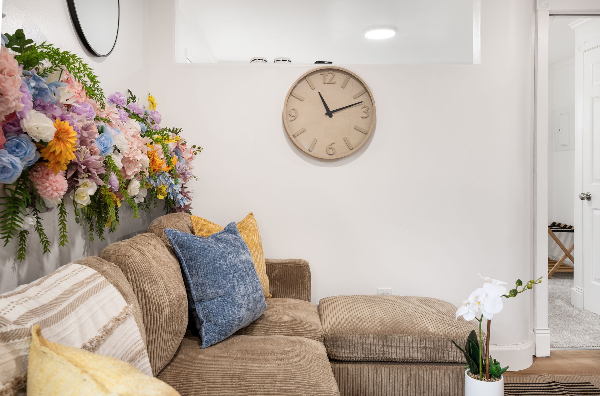 Cozy living room with beige sectional, blue pillows, flowers, and a wall clock above the couch