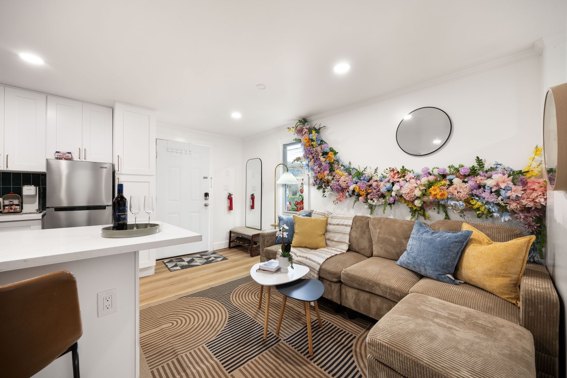 Bright living room with tan sofa, colorful floral wall decor, round mirror, and open kitchen counter