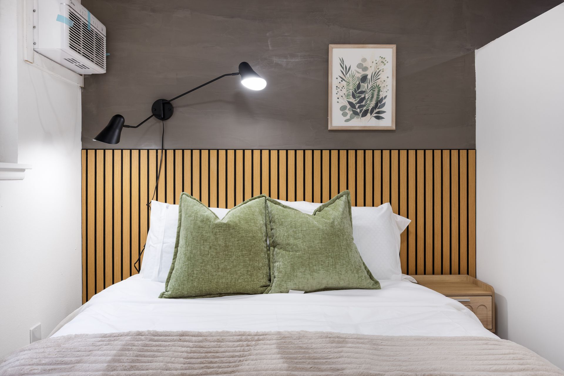 Modern bedroom with white bed, green pillows, wooden slat headboard, wall lamp, and framed plant print