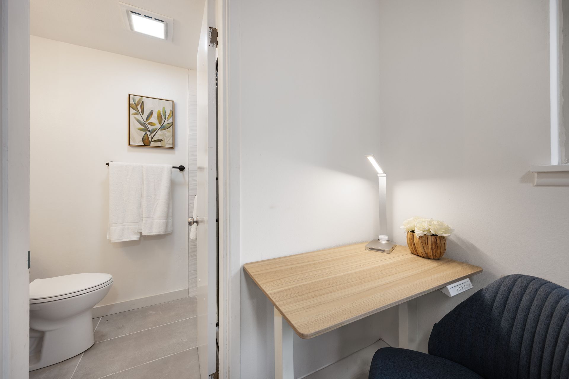 Small bathroom with toilet, towel rack, and a compact desk beside a chair under bright light.