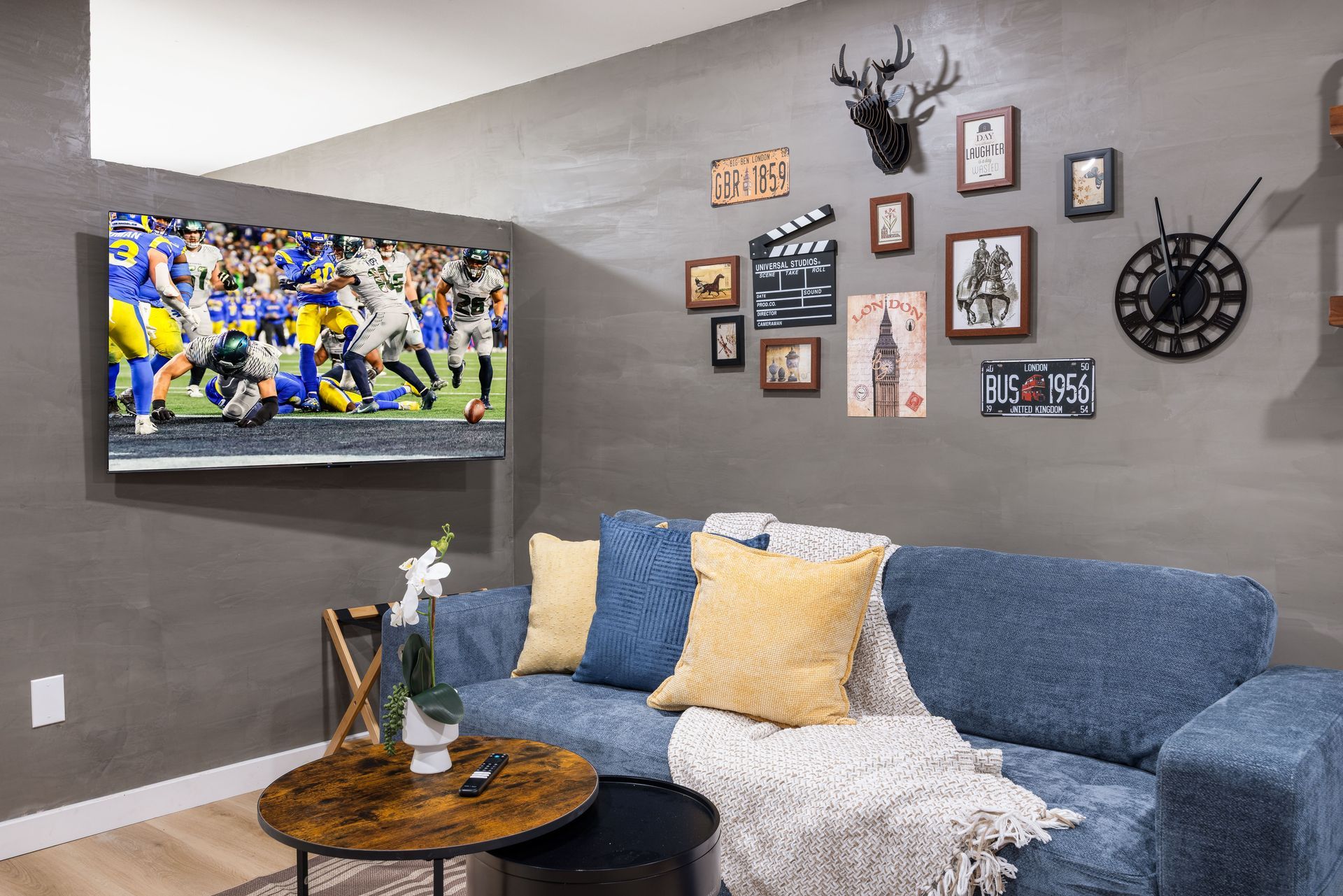 Cozy gray living room with wall-mounted TV showing a football game, blue sofa, and framed wall art.