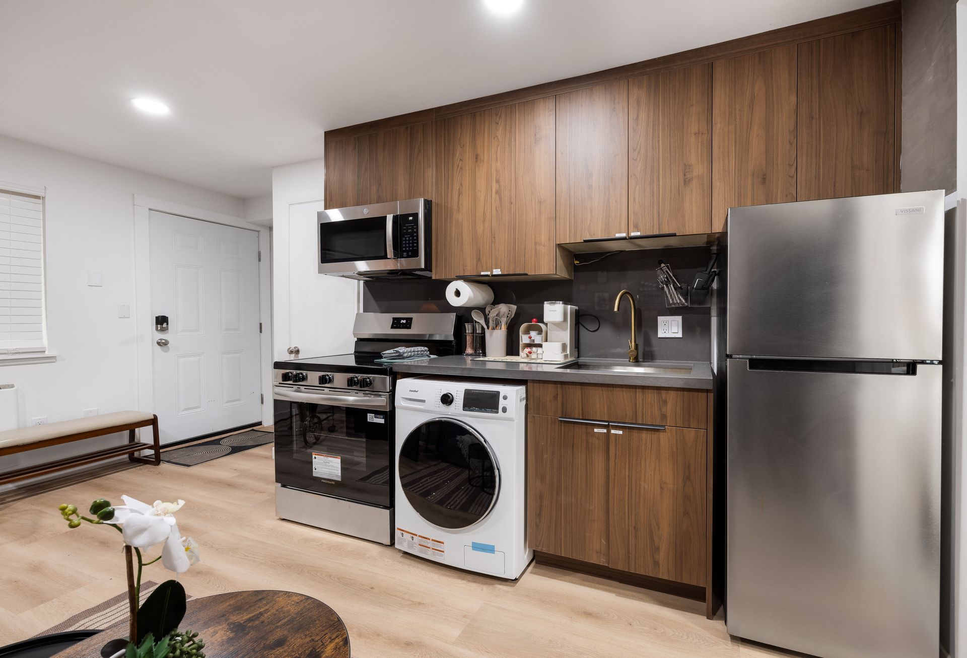 Modern apartment kitchen with wood cabinets, stainless appliances, and a dining bench by the entry.