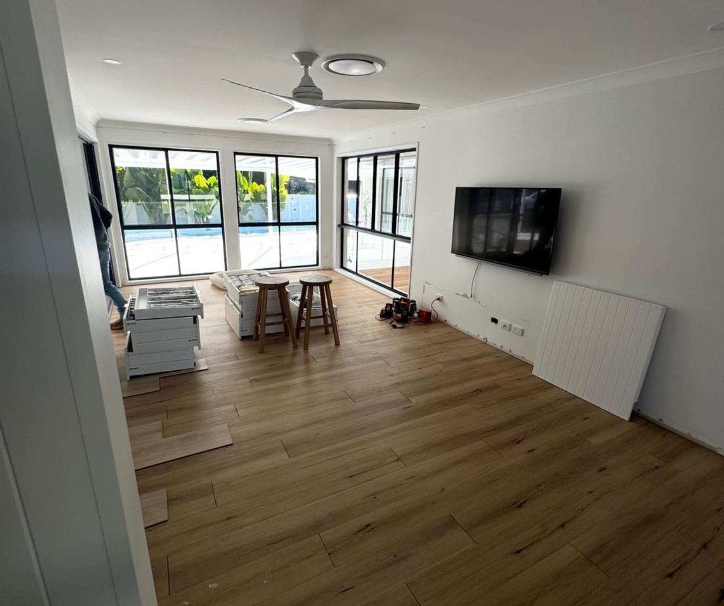 Bright modern living room with wood floors, wall-mounted TV, and large glass doors opening to a pool.