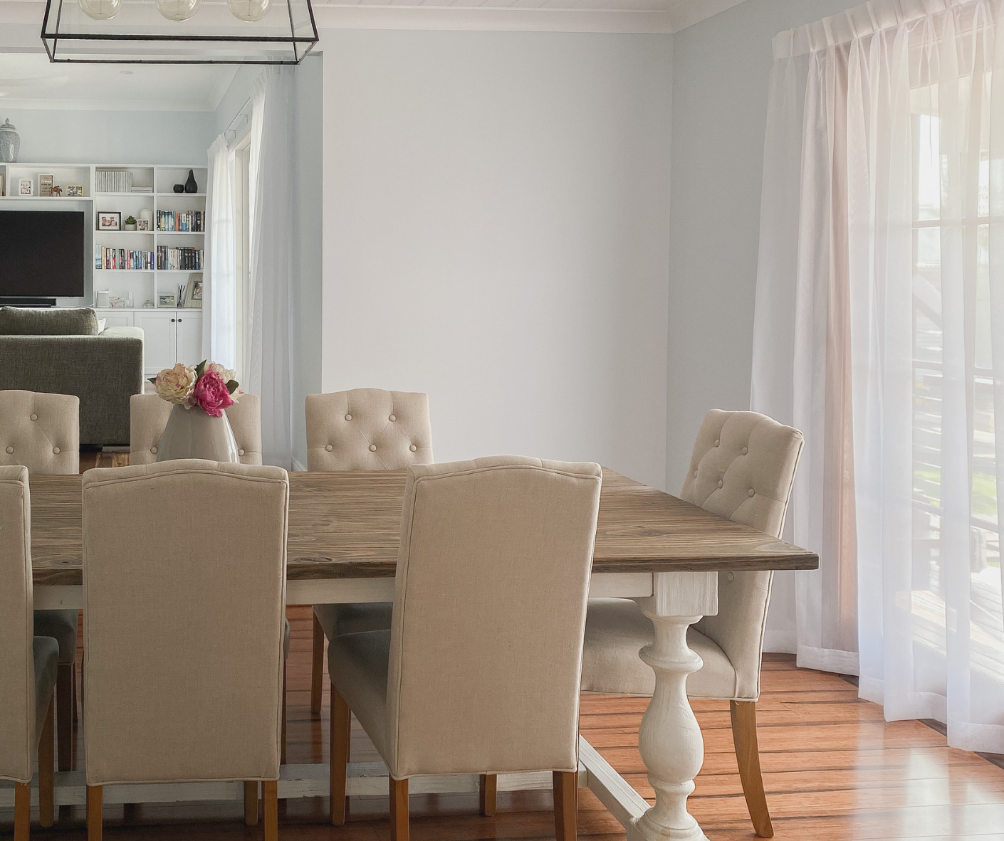 Bright dining room with beige chairs, a wooden table, and sheer curtains over a sunlit window.