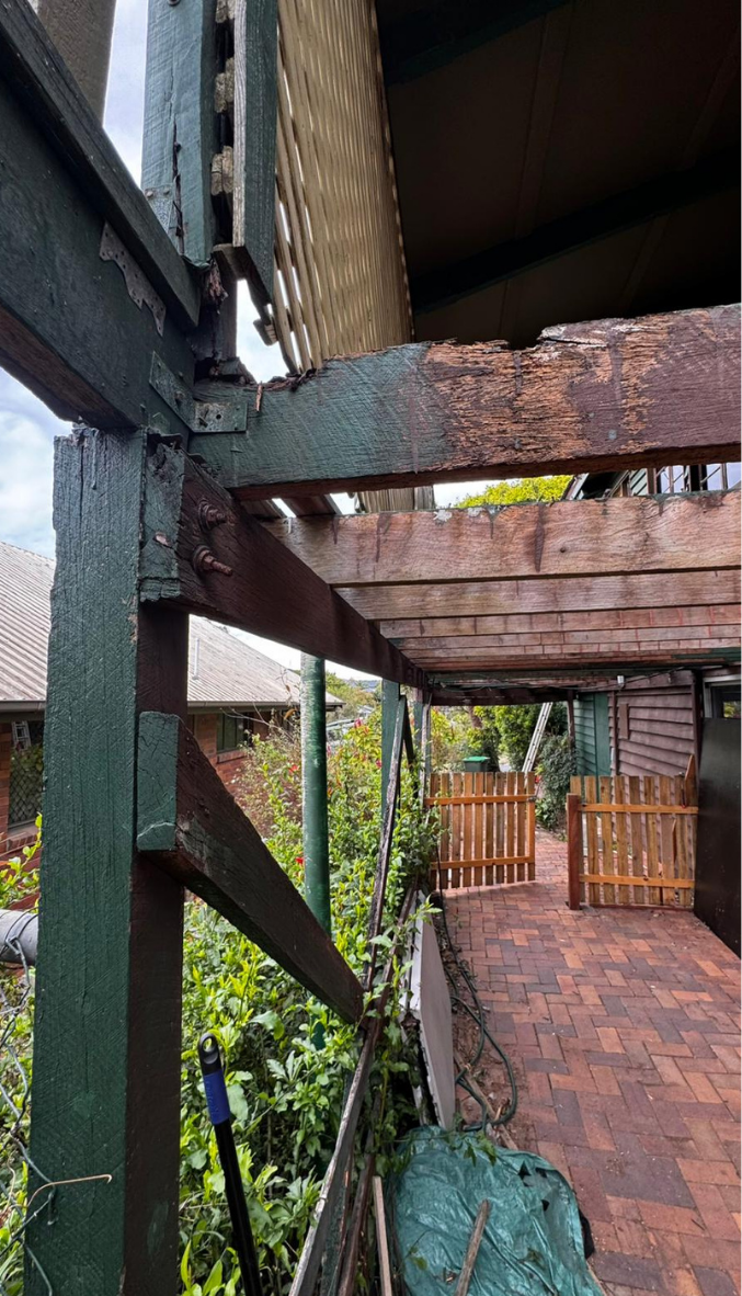 Rusty wooden beams and a brick walkway under a raised porch, with a fence and greenery beyond. Rusty wooden beams and a brick walkway under a raised porch, with a fence and greenery beyond.