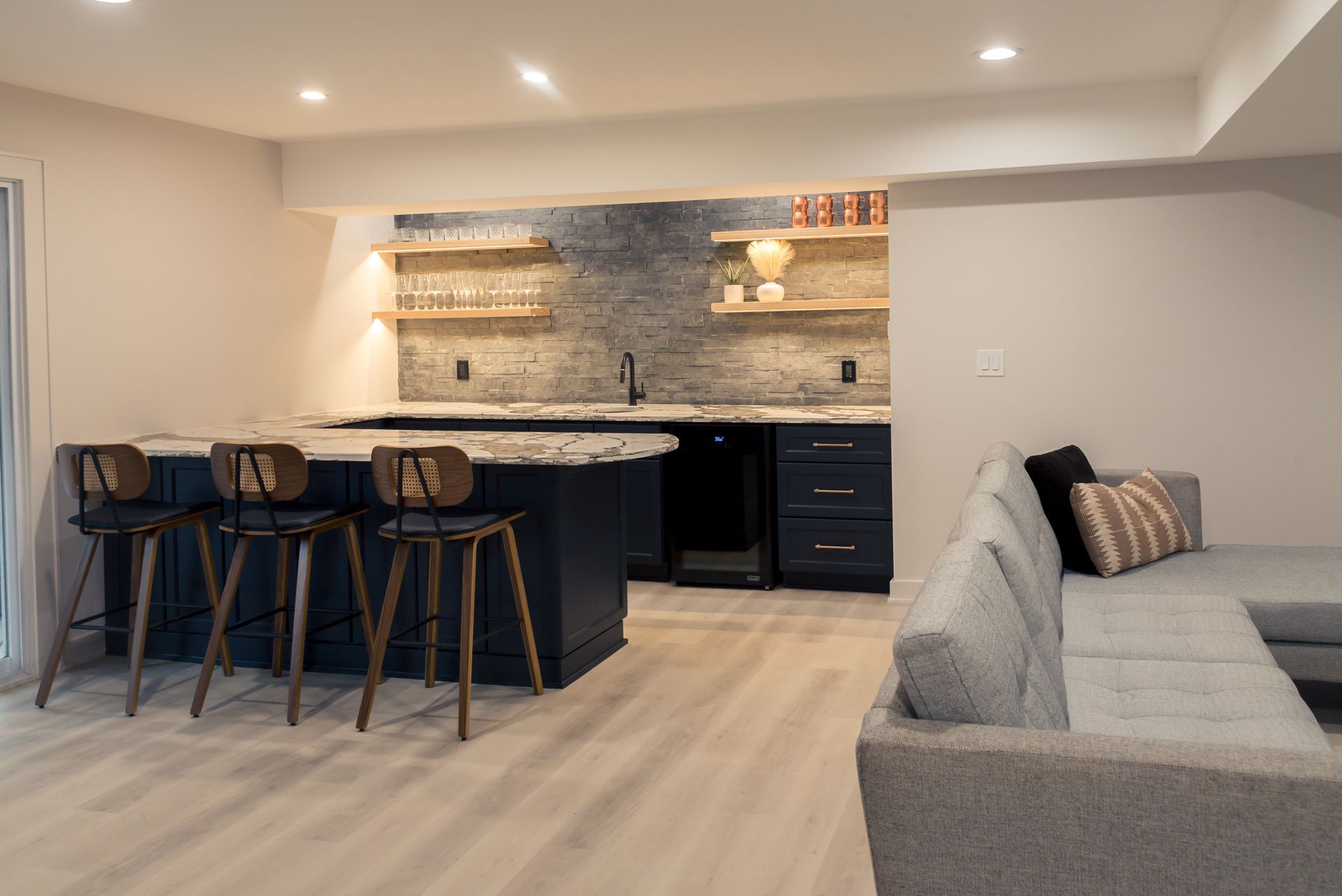 Modern basement kitchen and lounge with black island stools, gray sofa, and warm under-cabinet lighting
