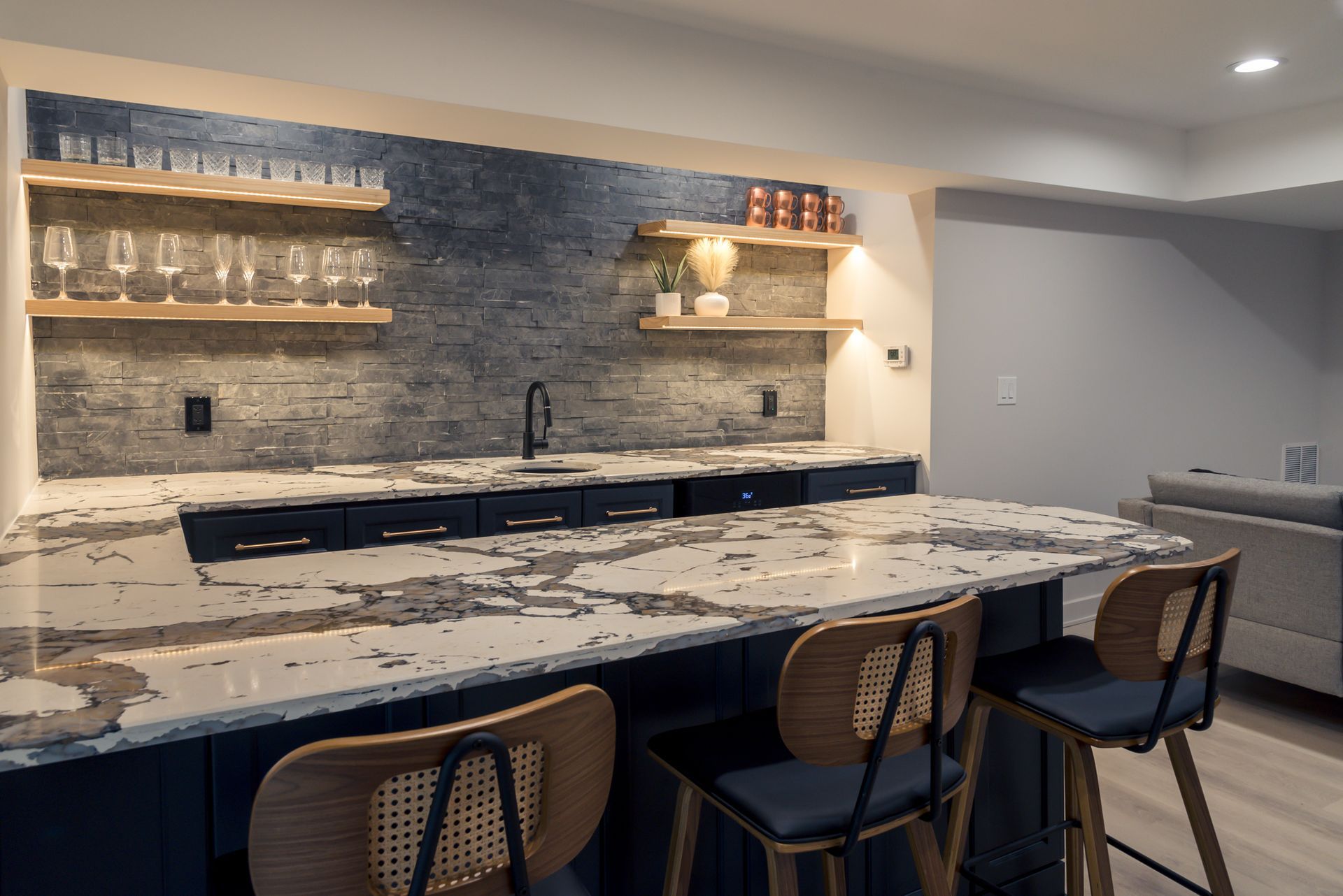 Modern kitchen with marble island, three woven stools, open shelving, and a black sink against a stone backsplash