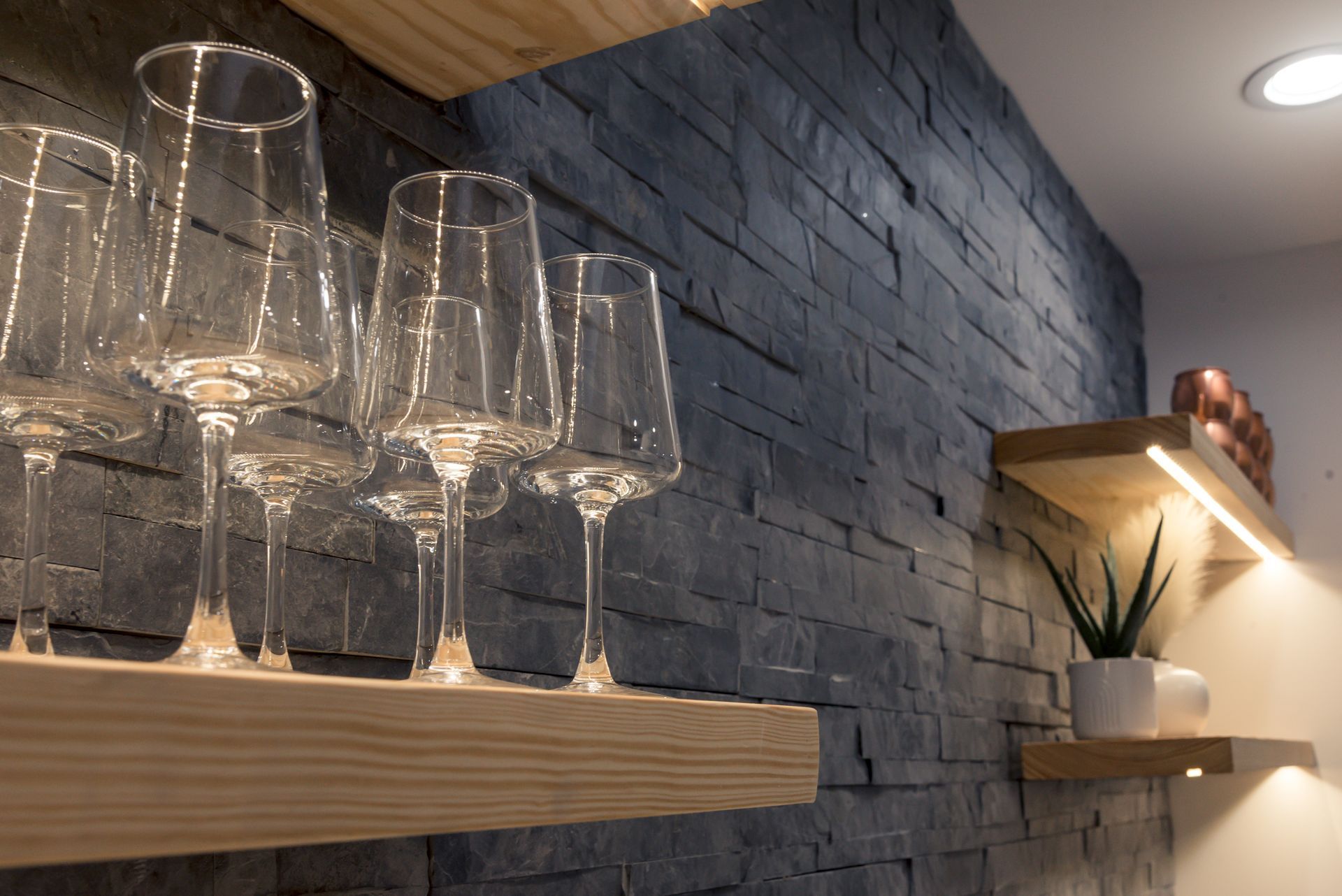 Wine glasses on a wooden shelf against a dark stone wall with warm accent lighting.